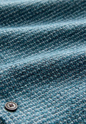 Close-up of textured blue fabric with a small dark button sewn on the lower left side.
