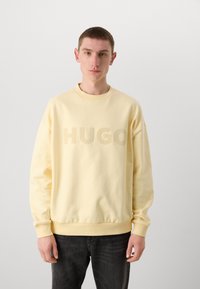 Sweatshirt amarelo com decote redondo e punhos canelados, apresentando um logótipo tonal "HUGO" na parte da frente, usado com jeans escuros.