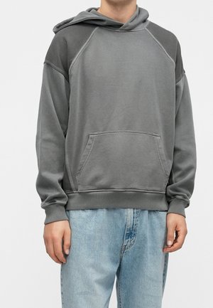 Person wearing a loose gray hoodie with a front pocket and blue faded jeans, standing against a plain background.
