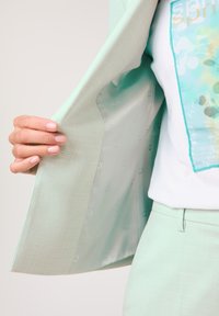 Minzgrüner Blazer aus strukturiertem Stoff mit glattem Innenfutter. Die Jacke ist offen und zeigt ein bedrucktes weißes T-Shirt mit bunten Akzenten.