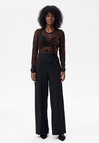 Vrouw met krullend haar, gekleed in zwarte high-waist broek met wijde pijpen en een doorschijnend zwart langarmshirt met geborduurde bloemen.