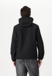 Casaco preto com capuz, feito de um tecido suave, apresentando um detalhe de costura nas costas e um bolso na manga esquerda. Combinado com jeans cinzentos.