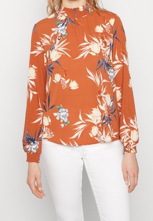 Vrouw draagt een roest-oranje blouse met hoge hals, wit-blauwe bloemenprint en gerimpelde manchetten, gecombineerd met witte broek.