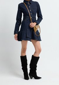 Femme portant une robe chemise en denim foncé avec un ourlet à volants, des bottes noires décontractées montant jusqu'aux genoux, et un petit sac bandoulière beige.