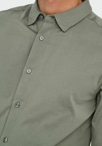 Chemise grise à manches longues en tissu doux, avec un col pointu et des coutures visibles. Comprend deux boutons à l'avant et une texture lisse.