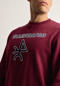 Sudadera burdeos hecha de una tela suave con un gran logotipo bordado que lleva el texto "VALECUATRO" y un diseño geométrico en azul marino.