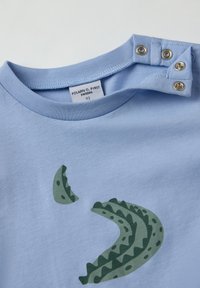 Vaaleansininen puuvillapaita, jossa on vihreä abstrakti kuvio. Olkapäillä olevat nepparit helpottavat pukemista. Tagissa lukee "Polarn O. Pyret."