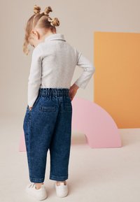 Maglietta grigia a maniche lunghe abbinata a pantaloni in denim blu con elastico in vita. Sneakers bianche. Lo sfondo presenta un arco rosa e un rettangolo arancione.