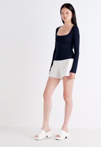 Un mannequin se tient devant un fond uni, portant un haut à manches longues bleu marine, un short blanc avec des bordures en œillet, et des mules blanches à talons.