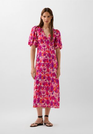 Robe portefeuille à fleurs avec des manches bouffantes dans des teintes vives de rose, rouge et violet. Dotée d'un décolleté en V et d'une coupe à la cheville. Portée avec des sandales noires.