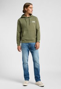 Felpa verde oliva con tasca frontale e cappuccio con coulisse, abbinata a jeans blu chiaro e sneaker beige. Presenta un logo bianco sul petto.