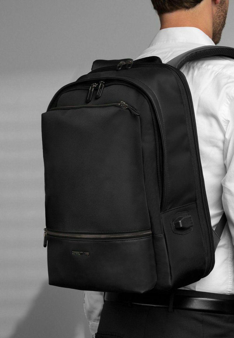 Muž v bílém tričku nese černý batoh s více přihrádkami, na kterém jsou viditelné taháčky zipů a polstrované ramenní popruhy.