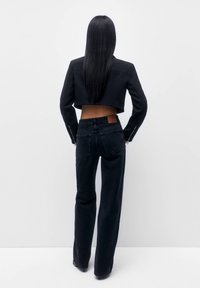 Woman with long straight black hair wearing a cropped black jacket and high-waisted black wide-leg jeans, standing against a white background.