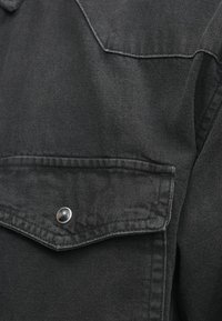 Czarna koszula z denimu z kołnierzykiem punktowym, przednią kieszenią z klapką i srebrnym zapięciem na zatrzask. Tkanina ma teksturę i lekko zużyty wygląd.