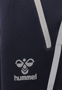 Marineblauer Sportstoff mit strukturierter Oberfläche, ausgestattet mit einem weißen Reißverschluss und einem auffälligen weißen Markenlogo "hummel" auf der unteren linken Seite.