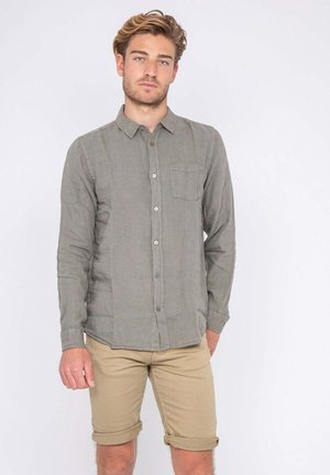 Jeune homme aux cheveux châtain clair portant une chemise grise à manches longues boutonnée et un short beige retroussé sur un fond uni.
