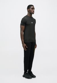 Melns īss piedurkņu t-krekls ar nelielu baltu logo uz krūtīm, kombinēts ar melnām sporta biksēm ar dzeltenu zīmolu un melniem sporta apaviem.