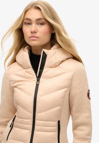 Kurtka puchowa z beżowym pikowanym przodem, czarnym zamkiem błyskawicznym i fakturowanymi, jasnobeżowymi rękawami. Posiada kaptur i naszywkę z logo na rękawie.