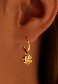 Boucle d'oreille en or avec un charme en trèfle à quatre feuilles, texture lisse et finition brillante, présentée sur une peau humaine.