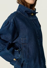 Denim jacket in dark blue, featuring a high collar, double buttons, loose sleeves, and a side pocket with contrasting stitching.