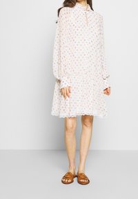 Robe à motifs floraux en tissu léger, avec un col volanté, des manches longues et une jupe à volants avec une fine dentelle. Des sandales marron complètent le look.