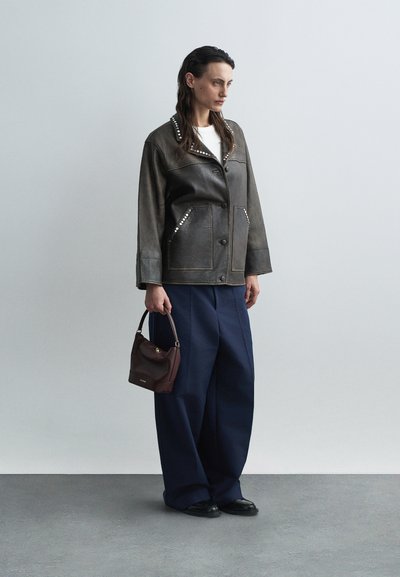 Braune Lederjacke mit Nietenkragen, weißes Hemd, marineblaue weite Hosen und eine kleine burgunderfarbene Handtasche. Schwarze Schuhe runden den Look ab.