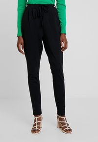 Zwarte broek met een ontspannen pasvorm, voorzien van een tailleband met trekkoord en zijzakken. Gepaard met lichtbruine sandalen met hoge hakken.