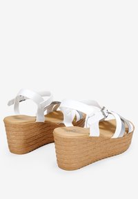 Sandales blanches à talon compensé avec une semelle en liège texturée, présentant des accents en métal et plusieurs sangles. Tige en cuir lisse et fermeture à boucle réglable.