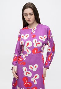 Μωβ φόρεμα με μεγάλα λουλουδάτα σχέδια σε κόκκινο και λευκό, μακρυά μανίκια, στρογγυλή λαιμόκοψη και απαλό, ελαφρύ ύφασμα.