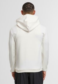 Felpa con cappuccio di colore bianco, caratterizzata da una finitura opaca liscia, pannelli laterali textured e polsini a coste. La vista posteriore mostra un design semplice senza loghi.