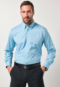 Camisa de botonadura azul claro con cuello estructurado, mangas largas y una textura suave, combinada con pantalones oscuros y un cinturón negro.