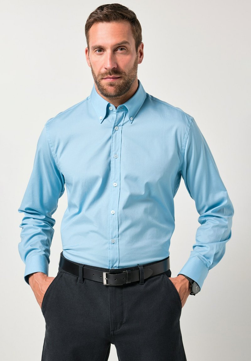 Camisa de botonadura azul claro con cuello estructurado, mangas largas y una textura suave, combinada con pantalones oscuros y un cinturón negro.