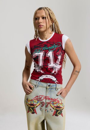 Personne aux longues tresses blondes portant un débardeur rouge et blanc Ed Hardy et un pantalon en denim clair avec des motifs de taureau graphiques et du texte.