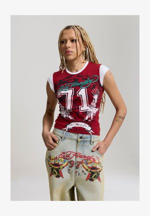 Personne aux longues tresses blondes portant un débardeur rouge et blanc Ed Hardy et un pantalon en denim clair avec des motifs de taureau graphiques et du texte.
