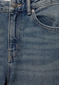 Sinivärvilised teksased, millel on nööpklapp, ees olevad taskud ja nähtavad õmblused. Kangas näitab kulunud tekstuuri ja kergeid pesu varieeruvusi.