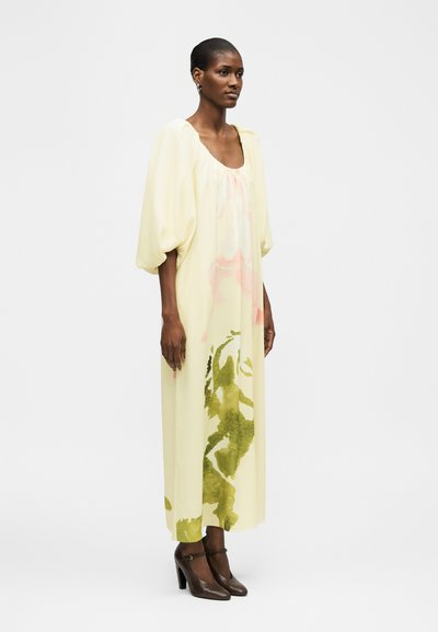 Γυναίκα που φοράει ένα μακρύ, ανοιχτό κίτρινο φόρεμα με φουσκωτά μανίκια και πράσινα λουλουδάτα σχέδια, σε συνδυασμό με σκούρα καφέ ψηλοτάκουνα παπούτσια.