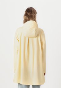 Chaqueta impermeable amarilla con capucha, de corte holgado y textura suave, que llega hasta la mitad de los muslos, sin herrajes ni estampados visibles.
