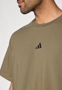 Λαδί βαμβακερό t-shirt με κοντά μανίκια, που διαθέτει μαύρο λογότυπο τριών ρίγων στο στήθος και εμφανείς λεπτομέρειες ραφής.