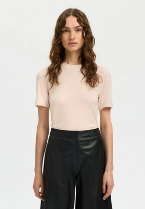 Femme aux cheveux longs et bouclés portant un t-shirt rose clair à manches courtes et un pantalon en cuir noir taille haute, jambes larges, se tenant devant un fond uni.