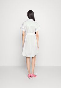 Abito camicia bianco con maniche corte, vita cintrata e orlo curvo. Abbinato a scarpe con tacco rosa con dettagli rotondi.