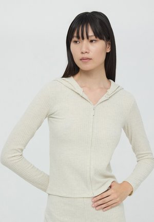Felpa con zip color beige chiaro, a coste, con un design aderente, maniche lunghe e cappuccio. Presenta una texture liscia e dettagli di cucitura sottili.