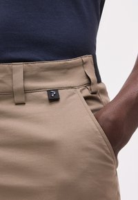 Κοντινό πλάνο ενός ατόμου που φοράει μπεζ παντελόνι με το χέρι στην τσέπη και μπλε σκούρο υφασμάτινο πουκάμισο που έχει μπει μέσα.