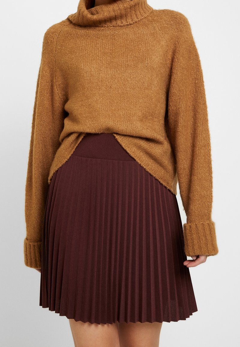 Pull à col roulé marron avec des poignets longs, associé à une jupe plissée taille haute de couleur bordeaux foncé, porté par une personne debout.