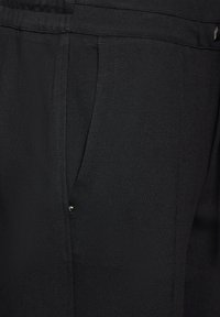 Svarta sportiga byxor med en slät textur, elastiskt midjeband, sidofaldar och en liten knappdetalj på framsidan.