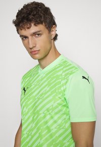 Zaļš īsās piedurknes sporta krekls ar ģeometrisku rakstu, nedaudz V veida kakla izgriezumu un melnu Puma emblēmu uz pleca. Gluda, viegla auduma.