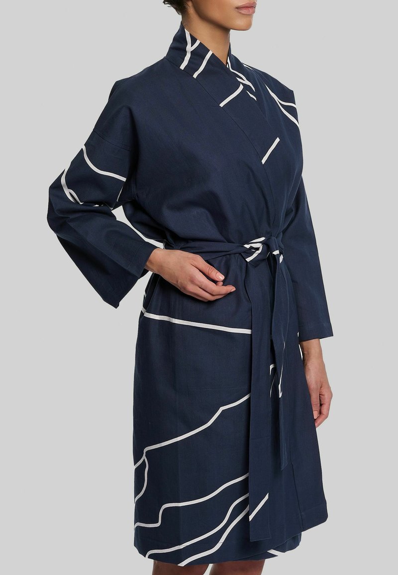 Femme portant une robe portefeuille bleu marine avec des lignes abstraites blanches, manches longues et une ceinture nouée à la taille sur un fond gris uni.