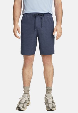 Man wearing navy blue drawstring shorts, light blue t-shirt, beige socks, and white and gray sneakers standing on a white background.