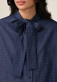 Blusa blu navy con pois bianchi, colletto a fiocco e chiusura a bottoni. Tessuto morbido con una trama liscia e un design strutturato.