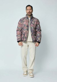 Doudoune avec un motif floral multicolore, poignets noirs et fermeture éclair. Portée par-dessus un pull aux tons neutres et un pantalon clair.
