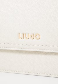 Carteira em imitação de couro branca com textura, apresentando o logo dourado "LIU·JO" e costura branca, destacando um design suave e minimalista.